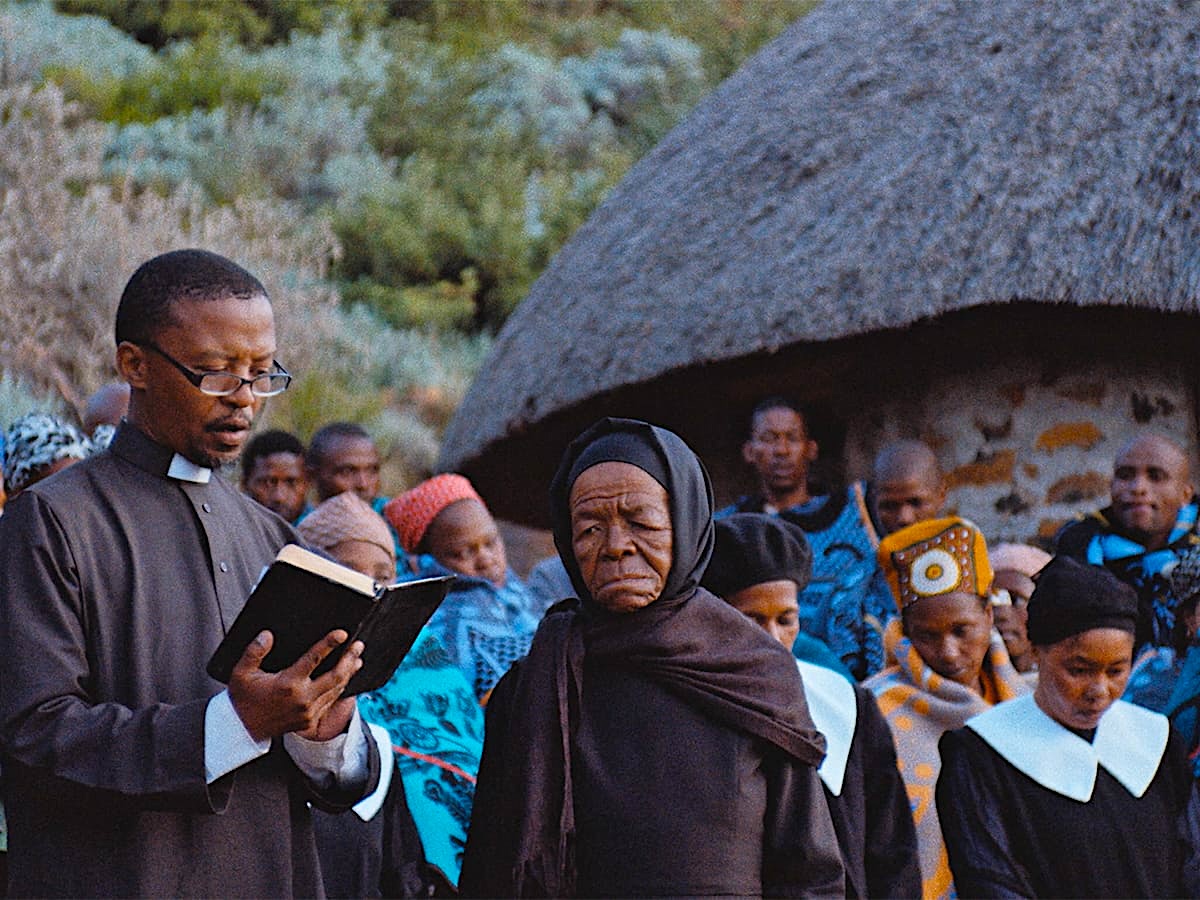 A priest reads from an open book beside an elderly woman dressed in black, while a gathered community stands outside a thatched-roof building during an outdoor ceremony.