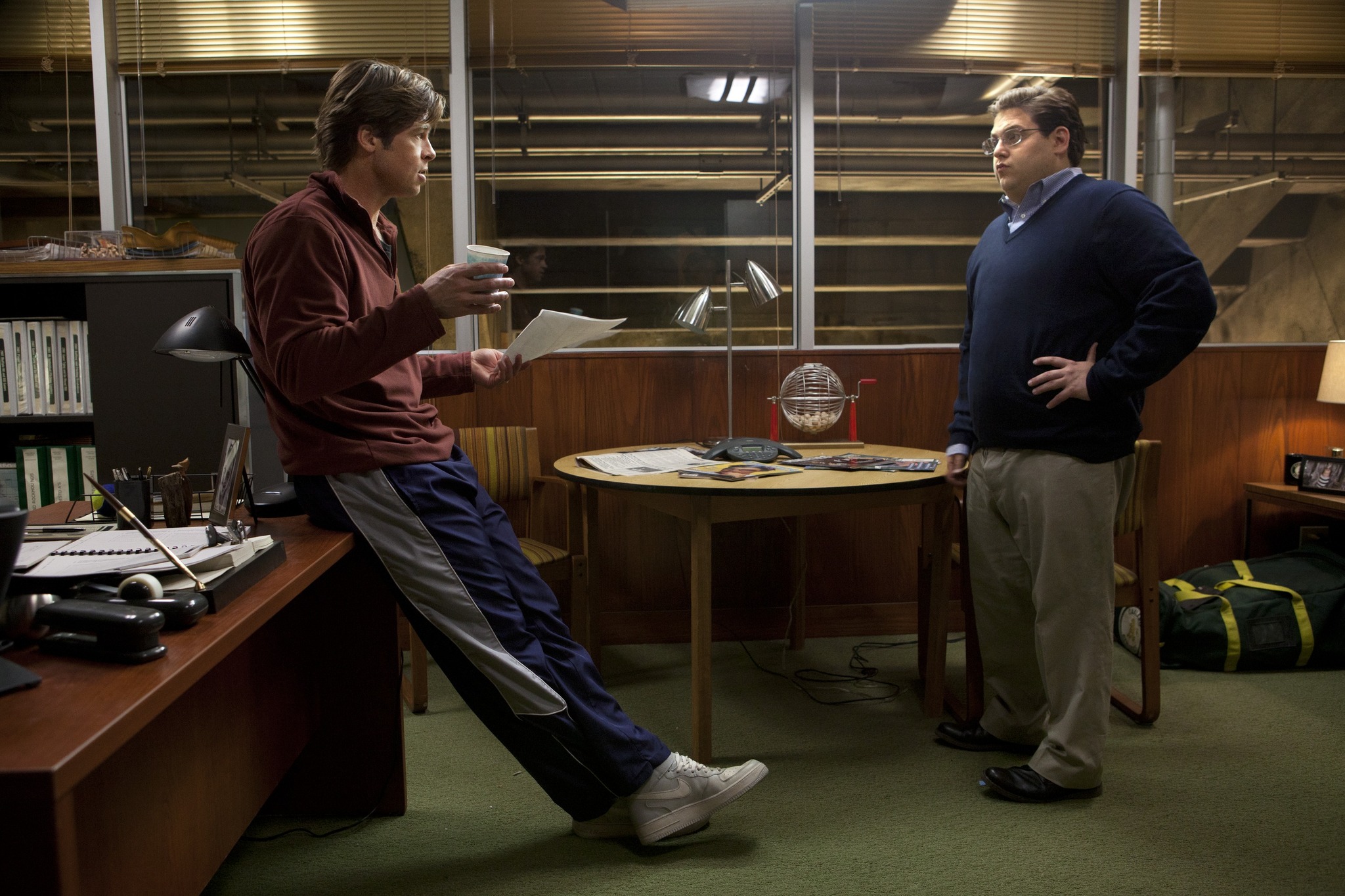 Two men stand in an office having a discussion, with one leaning against a desk holding papers and a coffee cup while the other stands with hands on his hips near a table.