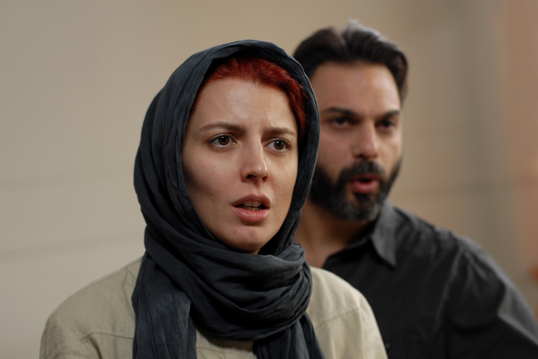 Close-up of two people indoors with concerned expressions. A person in a dark headscarf is in sharp focus in the foreground, while another person stands blurred behind them