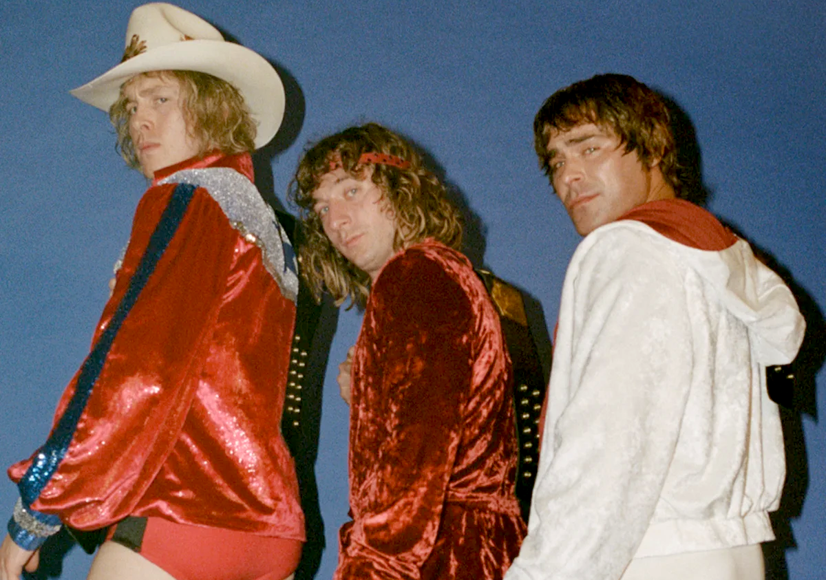 Three people in bright red and white performance costumes pose against a blue backdrop, looking over their shoulders toward the camera.
